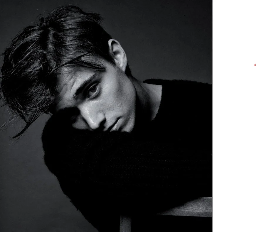 Black and white portrait of a young man with styled hair, resting his chin on his crossed arms, looking at the camera.