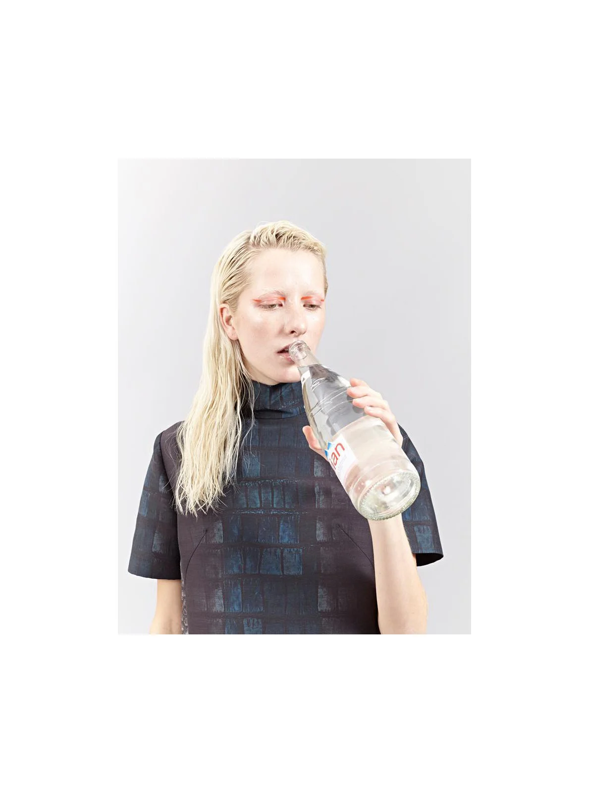 A person with blonde hair drinking water from a clear glass bottle labeled 'Voss' against a plain gray background.