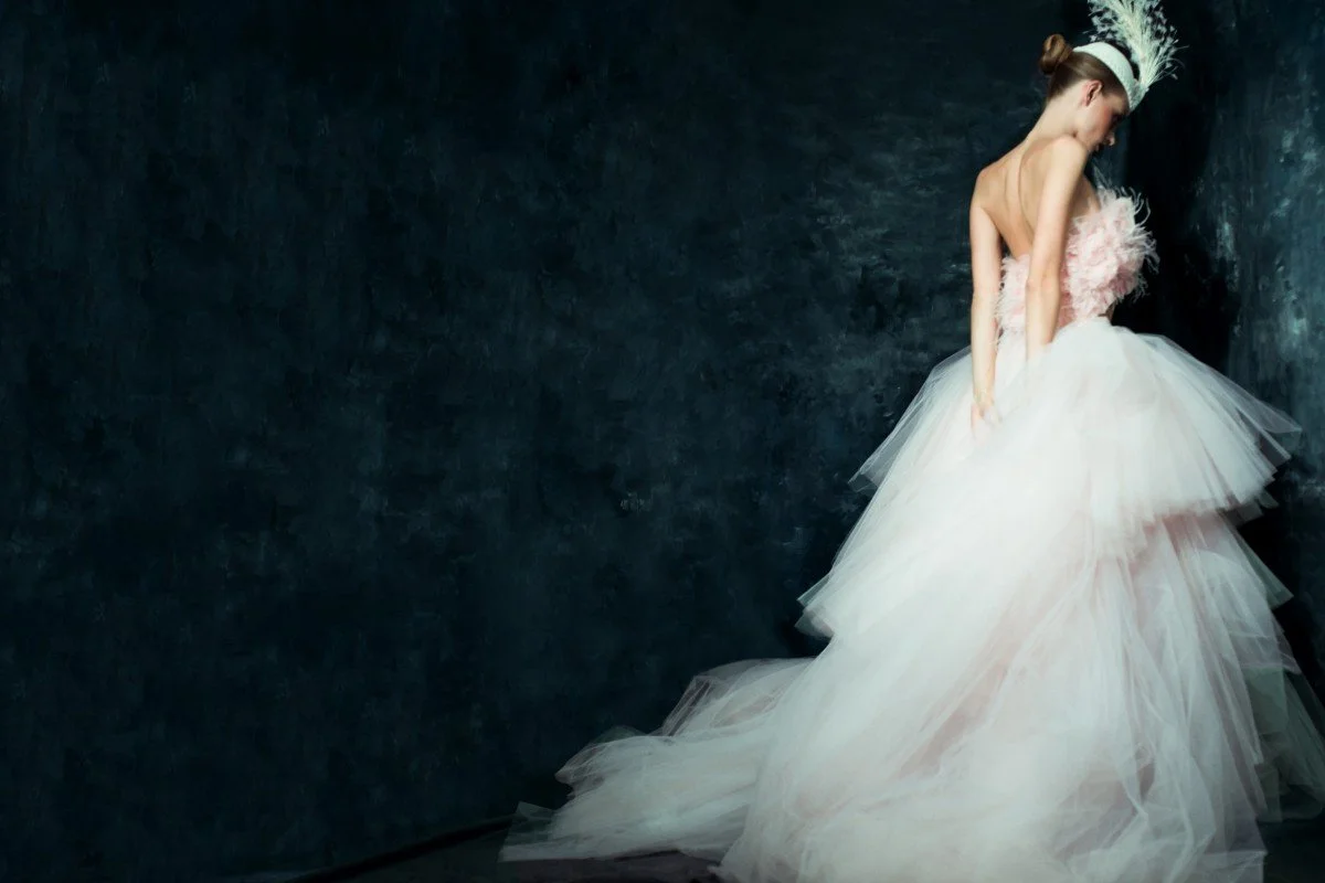 A woman in a pink and white wedding gown with a feathered headpiece standing against a dark wall.