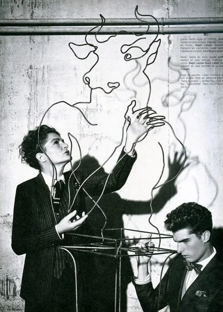 Black and white photo of a woman and a man creating a wire sculpture of a face with antlers on a wall. The woman is sewing and the man is holding wires.