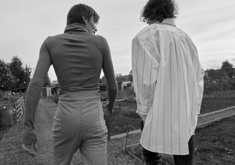 Two women walking together in an outdoor garden or farm setting, with trees and a cloudy sky in the background.