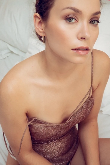 A woman with fair skin and brown hair styled in a bun, wearing a sparkly brown slip dress and small hoop earrings, sitting on a bed with white bedding.