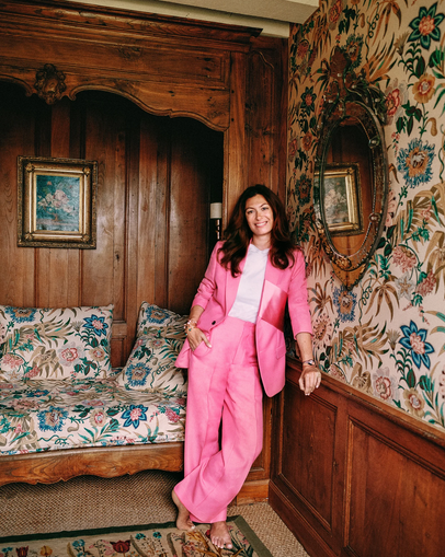 Woman in pink suit standing in a vintage room with floral wallpaper, wooden paneling, a mirror, and a floral-patterned sofa.