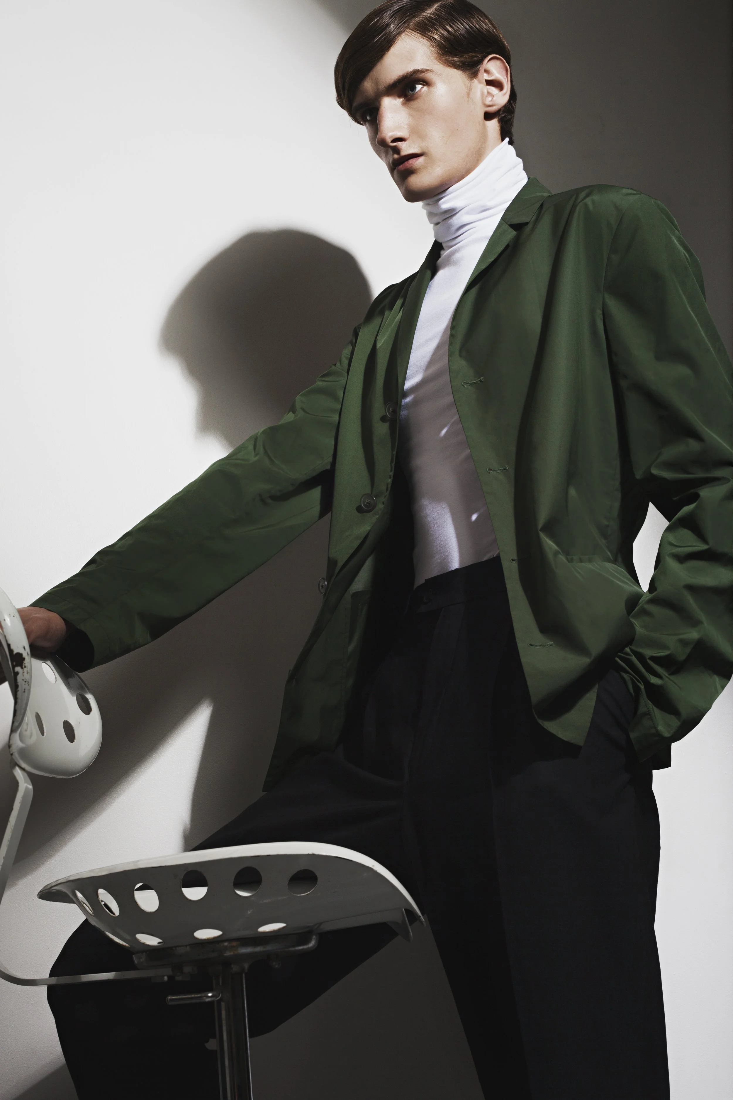 A young man with brown hair wearing a white turtleneck and a green jacket standing next to a white stool and a white sadle, with a shadow cast on a white wall.