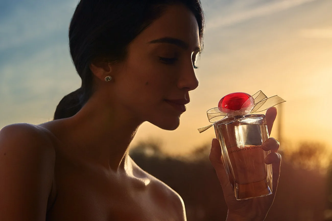 A woman with dark hair and earrings holding a perfume bottle with a red bow and cherry on top during sunset.