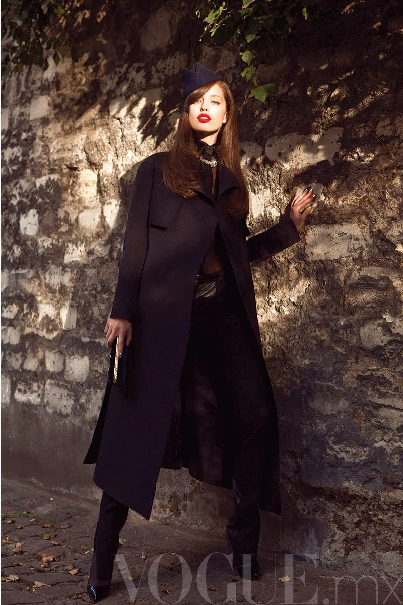 A woman with long brown hair wearing a dark navy coat, black pants, black shoes, and a navy hat stands against a stone wall, holding a small clutch purse.