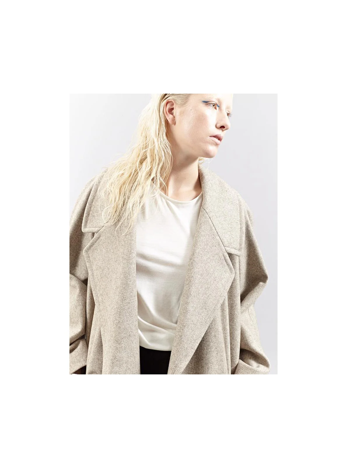 A woman with blonde hair wearing a beige coat over a white top, looking to her right against a plain light background.