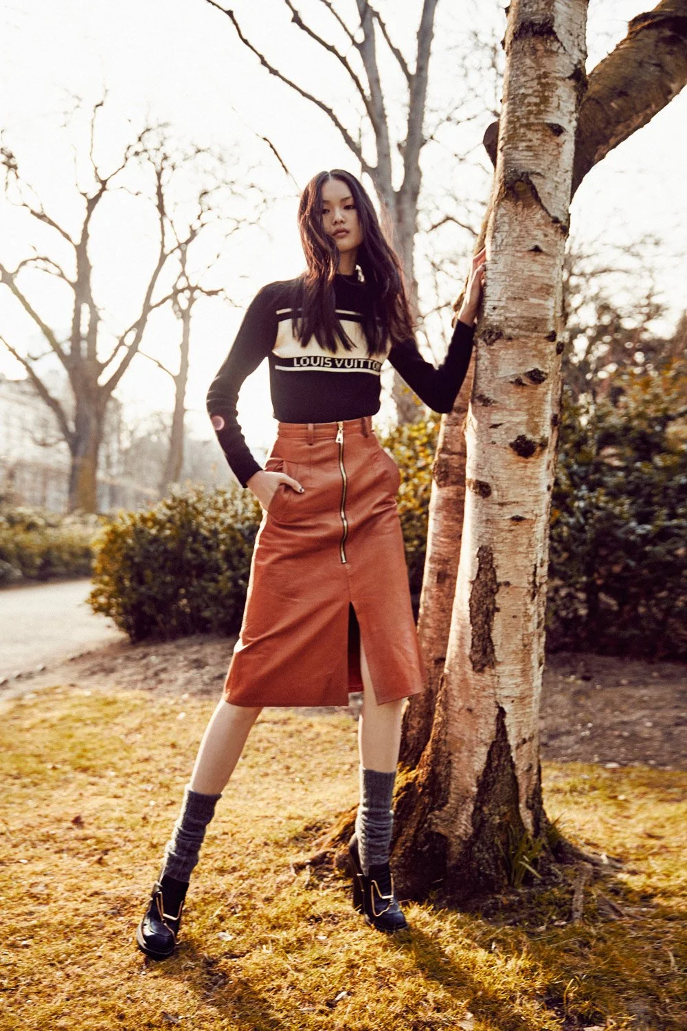 A woman standing outdoors in autumn, wearing a black Louis Vuitton sweater, a brown leather skirt with a front zipper and slit, gray socks, and black high-heeled shoes, leaning against a tree in a park.