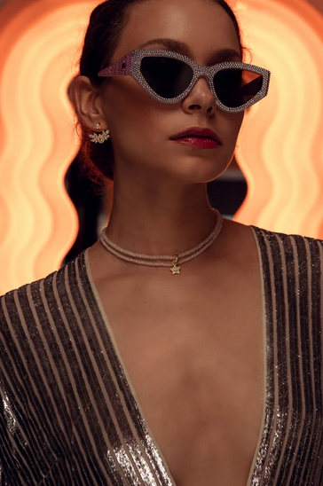 A woman wearing oversized, rhinestone-embellished sunglasses, a sparkly V-neck dress, star-shaped earrings, and a layered necklace, with a glowing orange background.