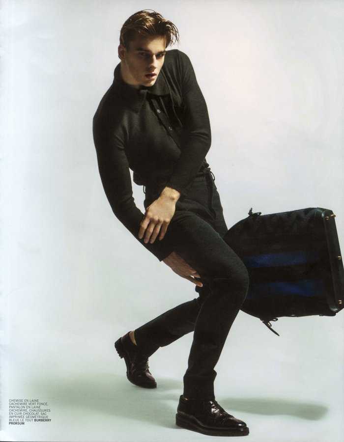 Young man in black clothing crouching sideways holding a large black bag, against a plain light background.