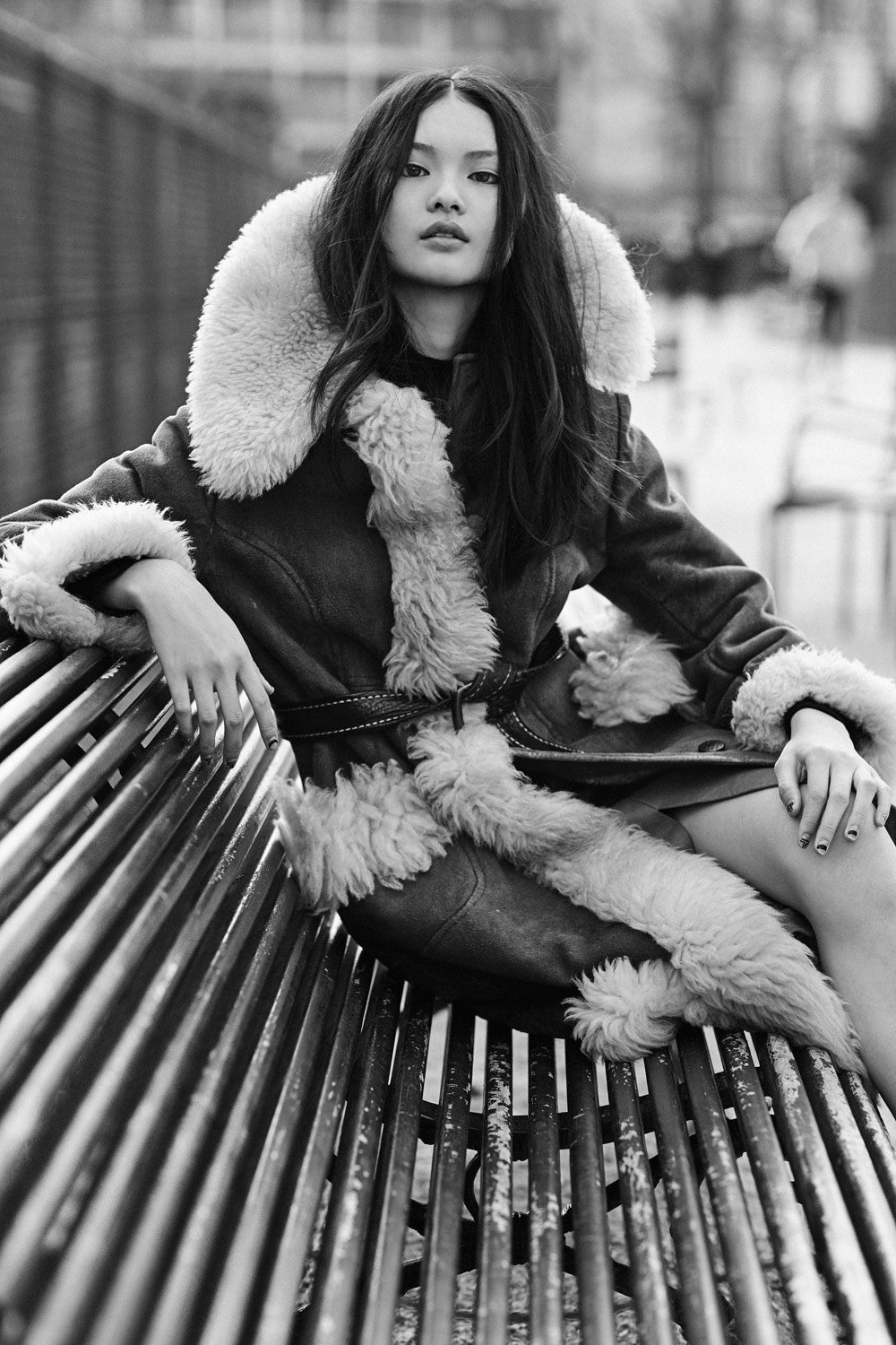 A young woman with long dark hair and light skin sitting on a park bench in black and white, wearing a dark jacket with fluffy lining. She has a serious expression and is looking directly at the camera, with her left hand resting on the bench and her