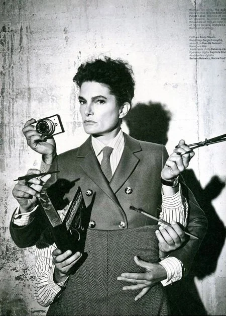 Black and white photo of a woman with short curly hair, dressed in a blazer, holding a camera. Multiple arms with various objects including a paintbrush, a pointer, and a notebook surround her, creating a surreal, multi-armed effect against a texture