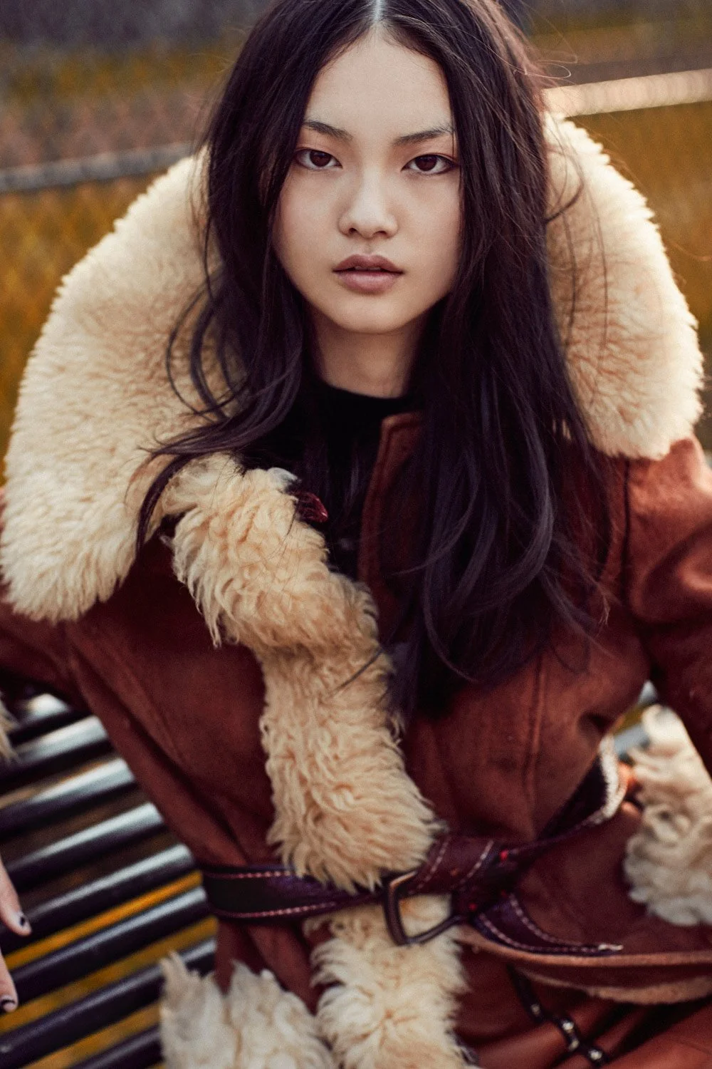 A woman with long dark hair and a serious expression wearing a brown shearling-lined jacket, sitting outdoors on a park bench with autumn foliage in the background.