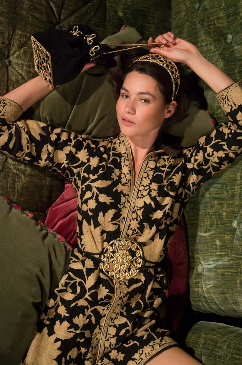 A woman with dark hair reclining on plush green and red cushions, wearing a black and gold floral embroidered dress with a large ornate gold belt and matching headband, holding a gold twig in her right hand.