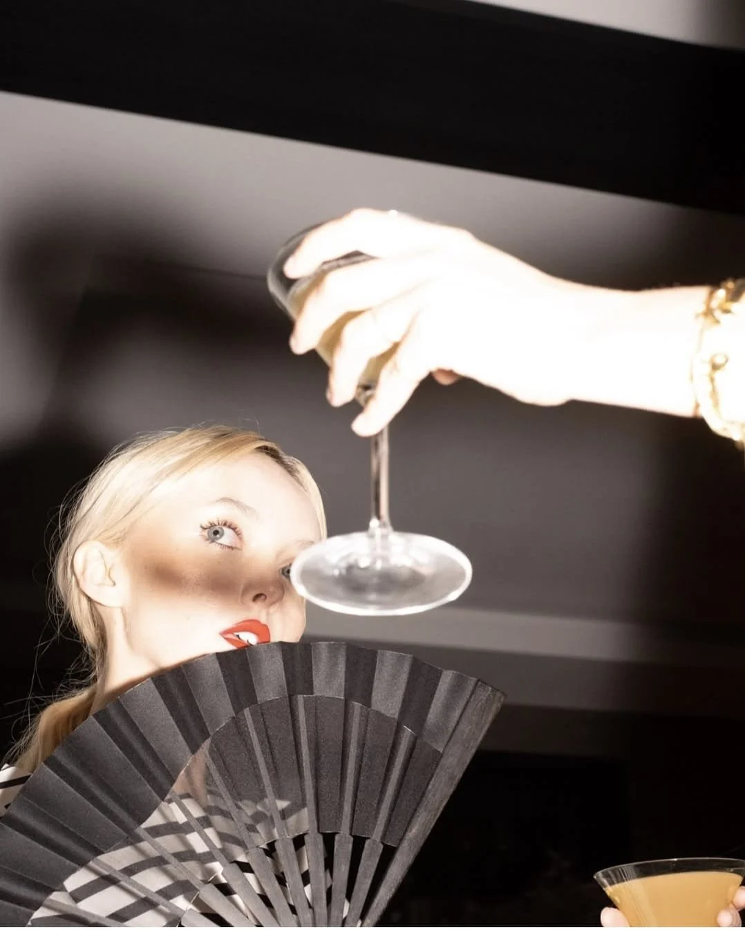 A woman with blonde hair and red lipstick holding a black fan partially covering her face, with a person above her holding a glass of wine.