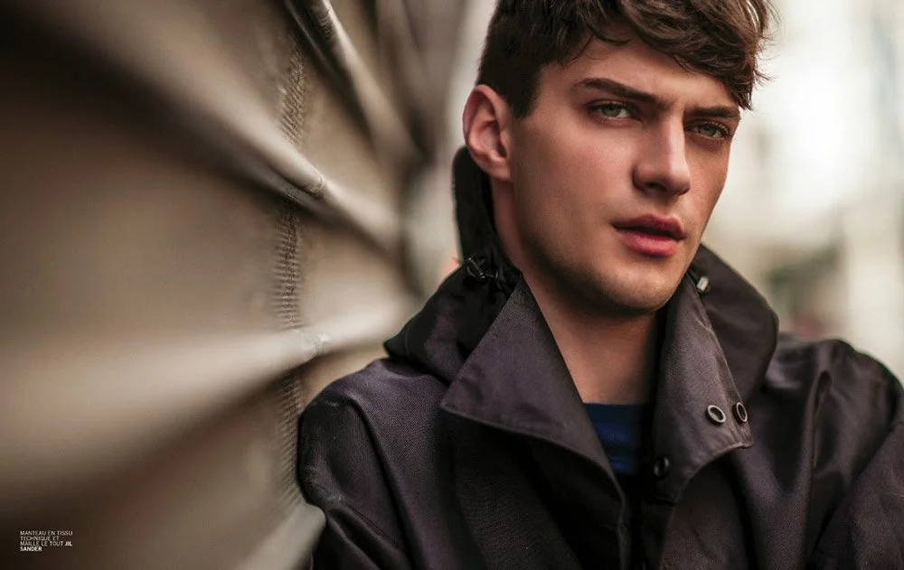 A young man with brown hair and blue eyes, wearing a black jacket, leaning against a metallic wall outdoors.