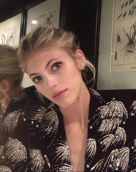 A woman with blonde hair styled in an updo, wearing a black sequined top, sitting in front of a mirror and wall decorated with framed botanical prints.