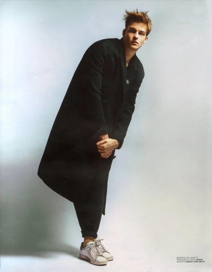 Young man with tousled hair wears a long black coat, black pants, and white sneakers, standing against a plain light background.