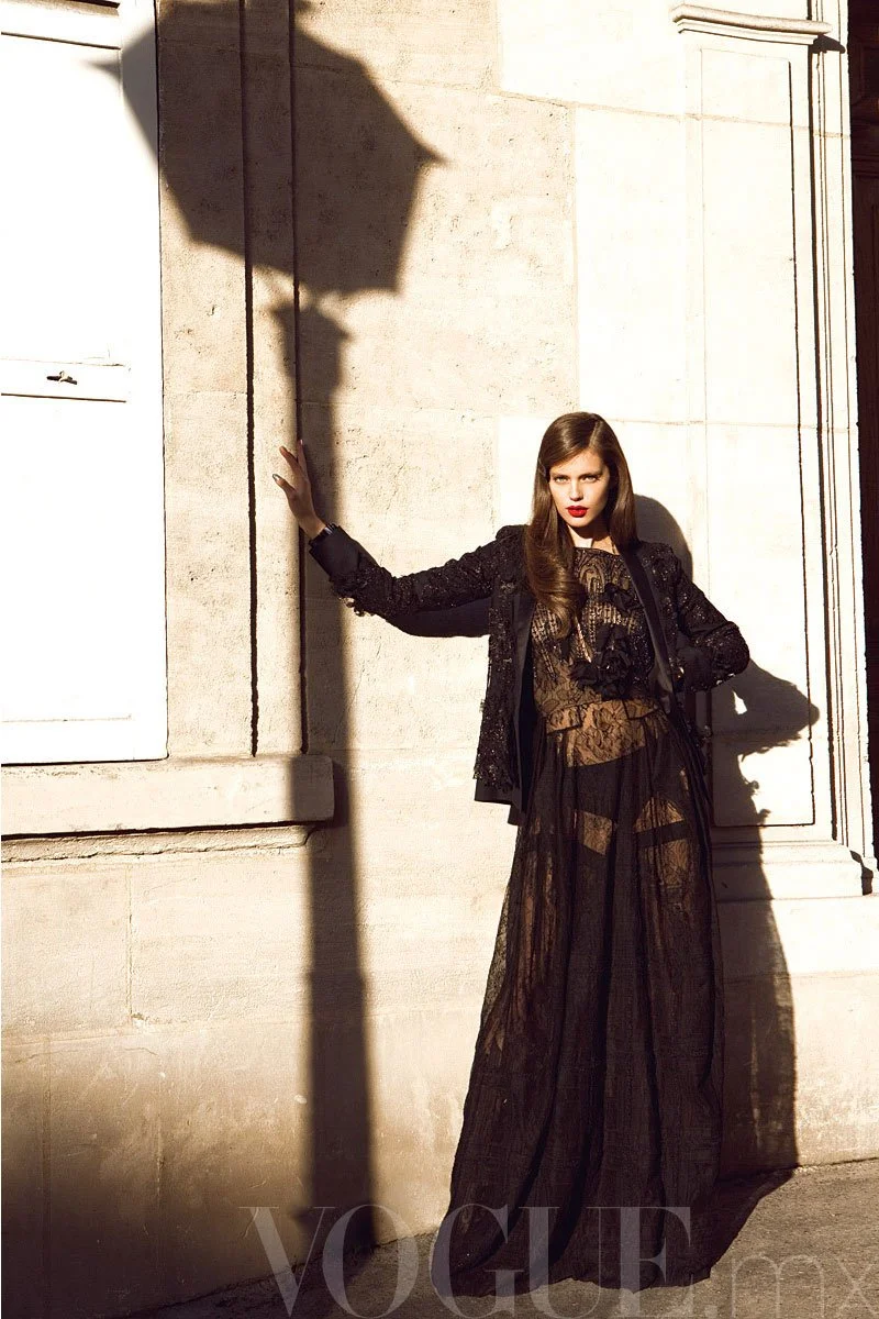 A woman in black sheer lace dress and black sequined jacket leaning against a stone wall at sunset, with her hand touching the wall and a shadow of a street lamp cast on the wall.