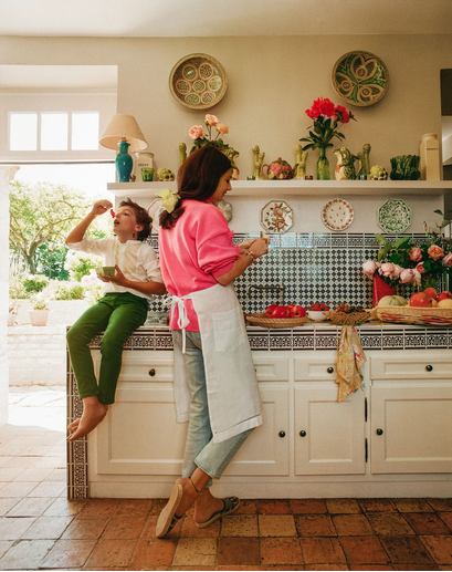 A woman and a child in a bright kitchen with various decorative plates and flowers, preparing or eating food.