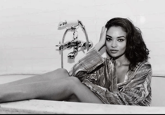 A woman lying in a bathtub, resting her head on her hand, with a relaxed expression, wearing a loose shirt.