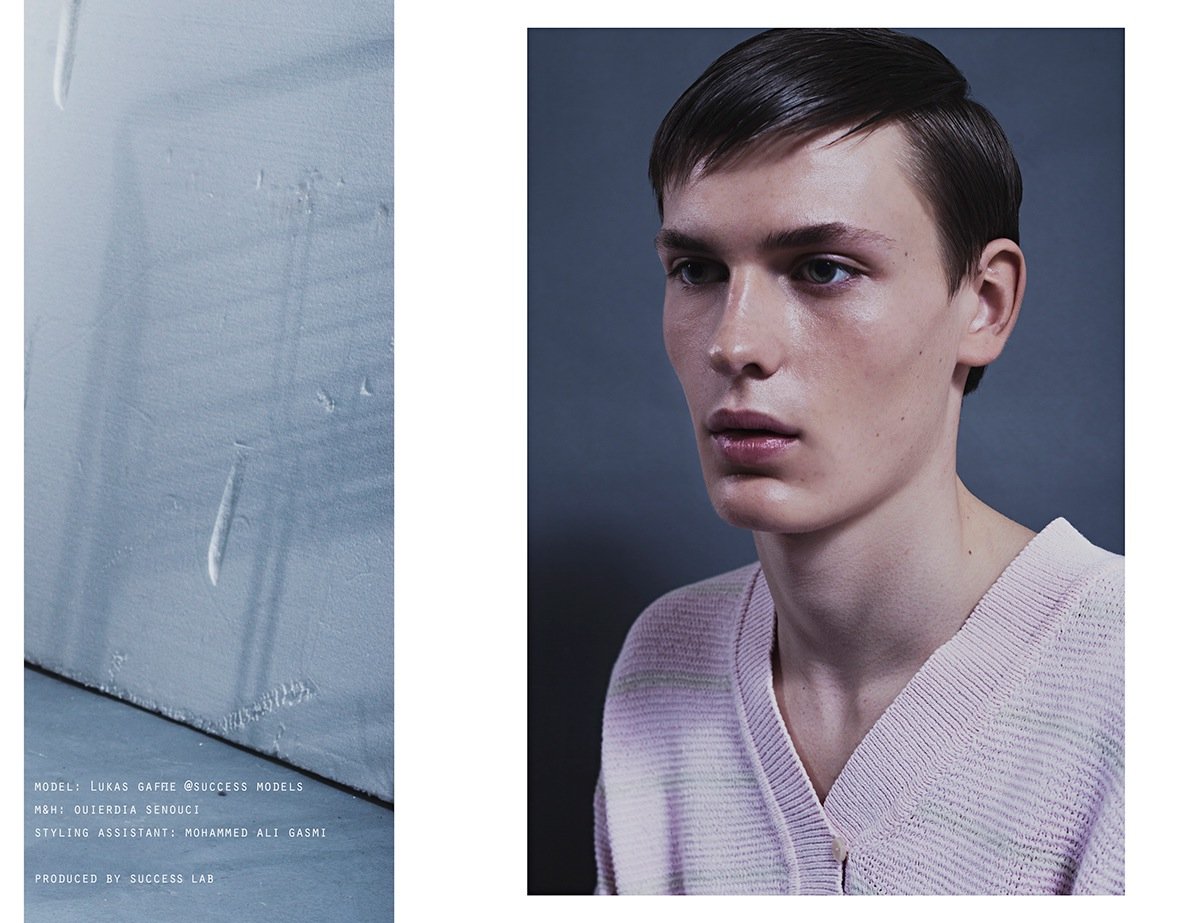 A portrait of a young man with short dark hair wearing a light pink knit sweater against a dark background. Beside him, there is a textured wall with text indicating this is a fashion or modeling shoot, featuring a photographer, stylist, and producti