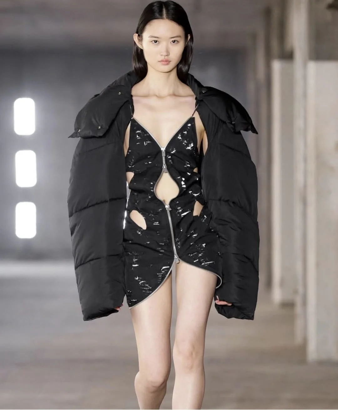 Fashion model walking in an industrial setting, wearing a black splattered zip-up dress with cutouts and a black puffy jacket draped over her shoulders.