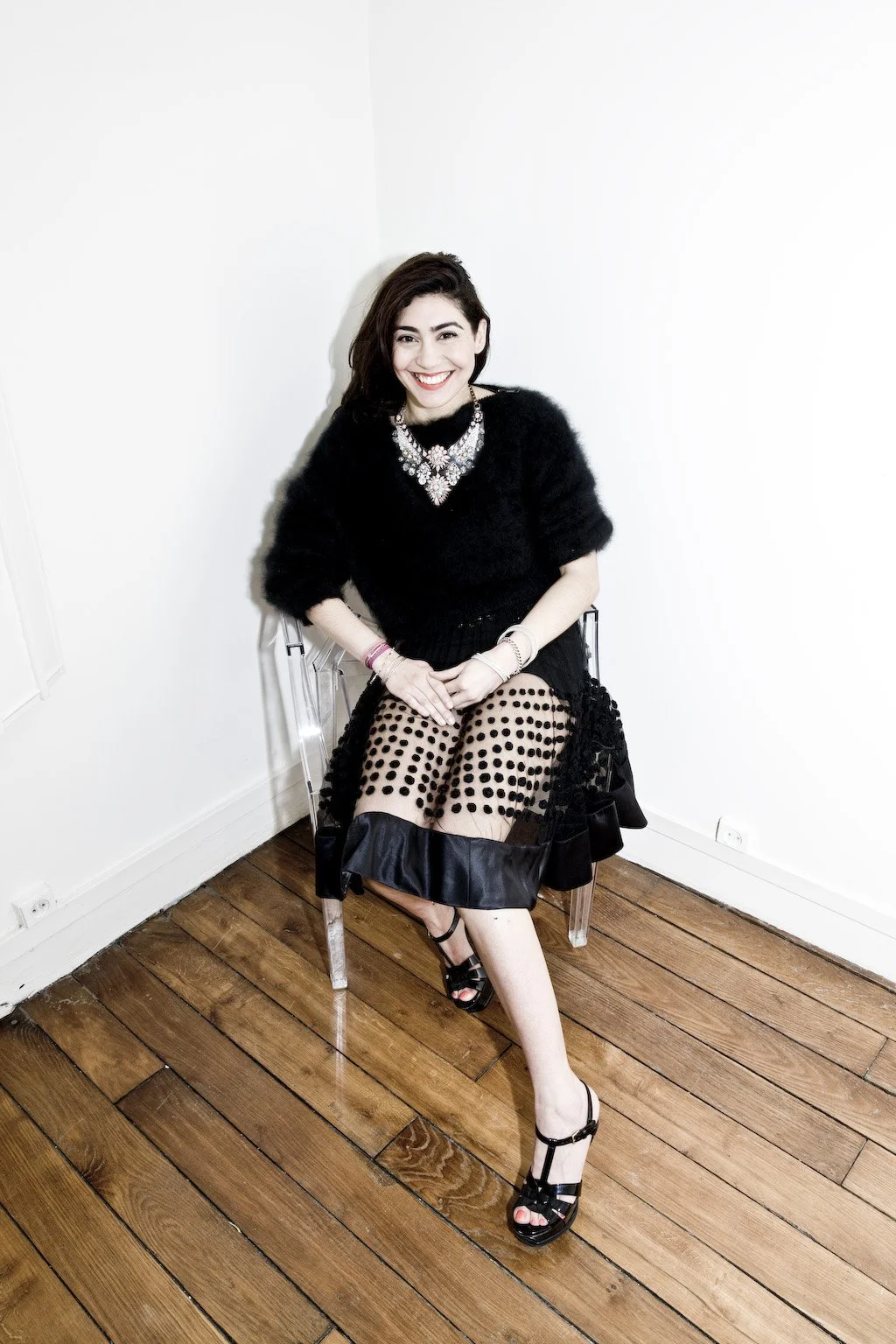 A woman with dark hair and a bright smile, dressed in an elegant black outfit, sitting on a clear acrylic chair in a minimalistic room with wooden floors and white walls.