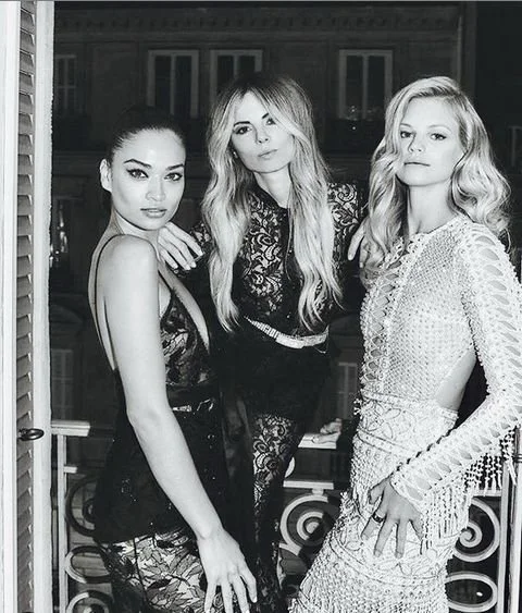 Three women posing together on a balcony at night, dressed in stylish outfits.