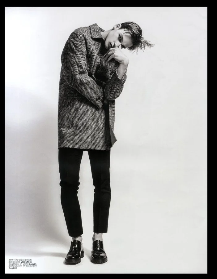 Black and white photo of a tall person wearing a herringbone patterned coat, black pants, and shiny black shoes, standing against a plain light background, with one hand near their face and eyes closed.