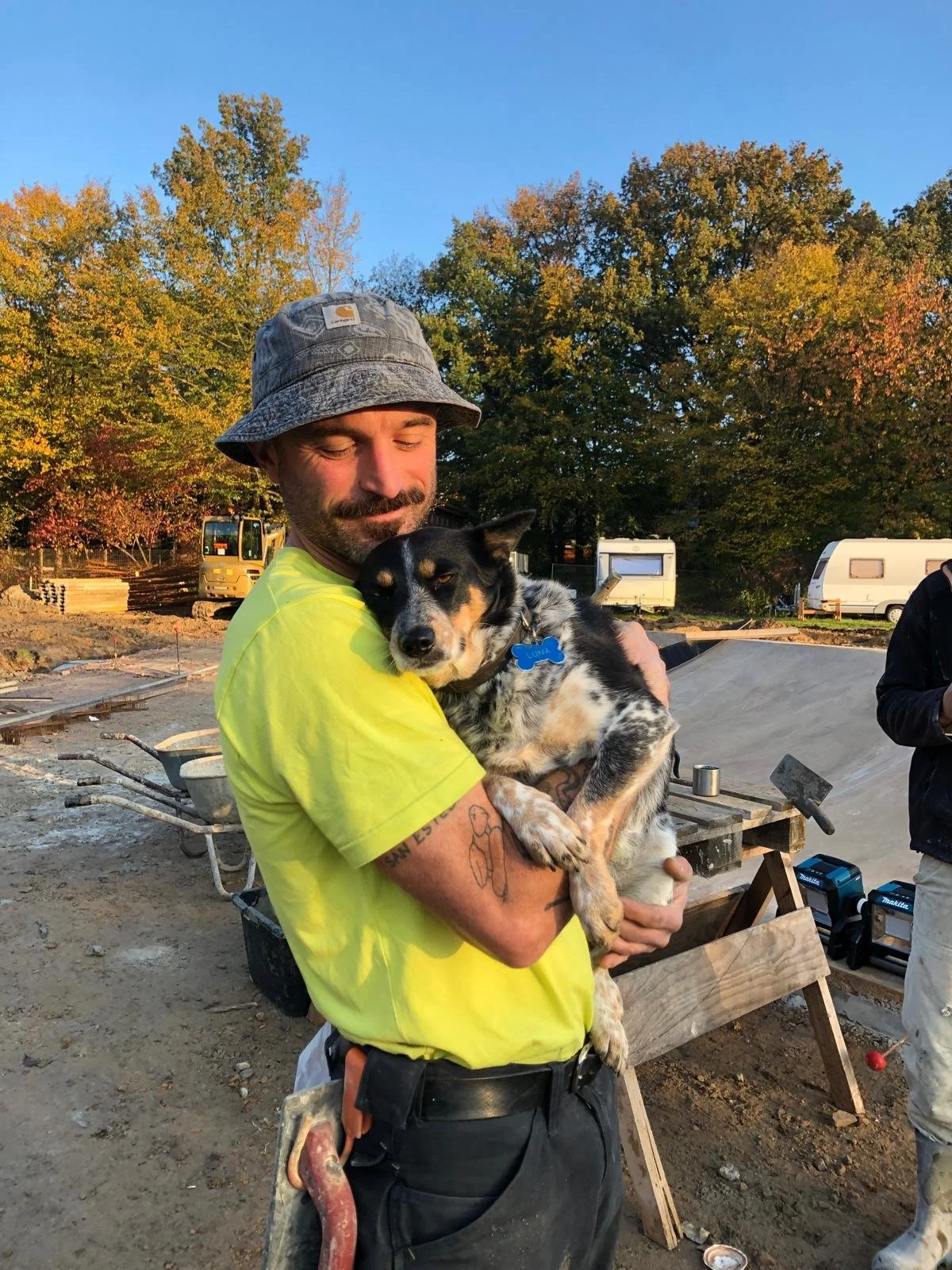 A man with a beard and wearing a gray bucket hat and a yellow shirt holding a black, white, and tan dog at a construction site during autumn. Trees with colorful leaves and trailers are visible in the background.