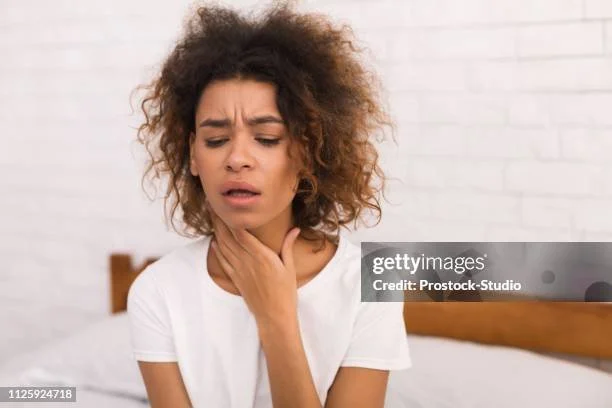 Una donna con capelli riccioli, vestita con una maglietta bianca, che si tiene la gola con un'espressione di dolore o disagio.