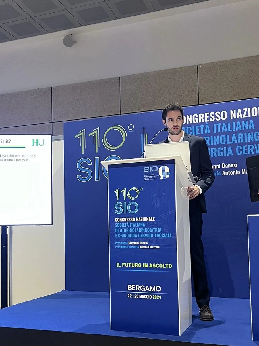 Uomo in piedi dietro un podio durante un congresso, con uno schermo a sinistra che mostra un diagramma, e sfondo con banner blu del congresso medico.