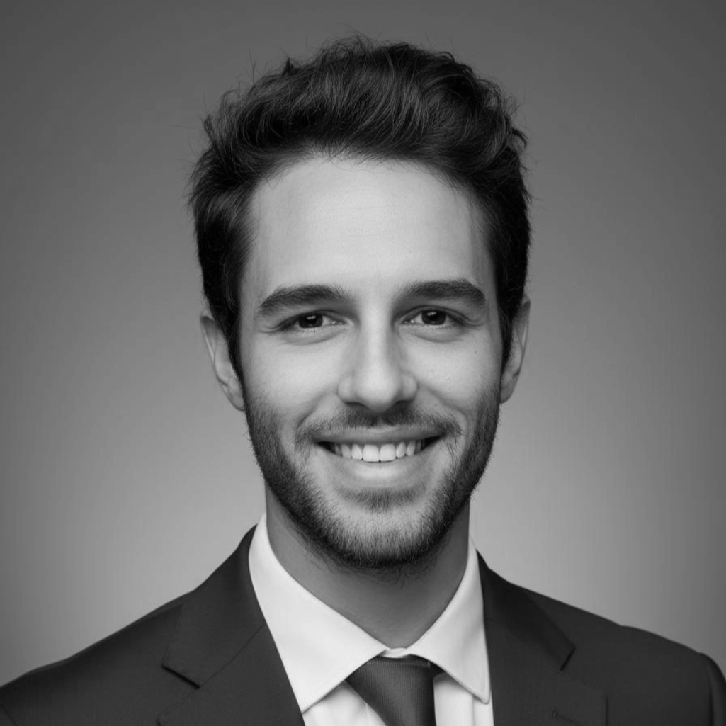 Foto in bianco e nero di un giovane uomo sorridente, con capelli scuri e barba leggera, indossa un abito con cravatta, sfondo sfocato.