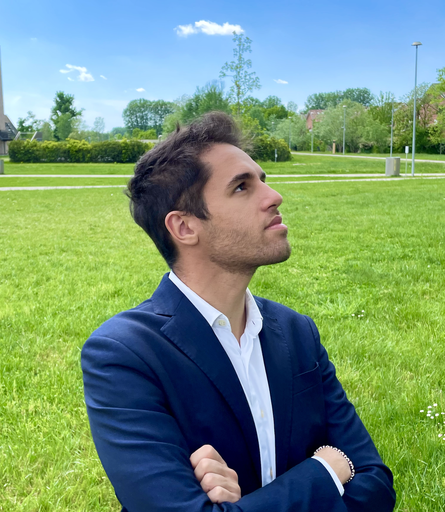 Uomo in giacca blu e camicia bianca con braccia incrociate davanti a un prato verde con alberi e luce del giorno