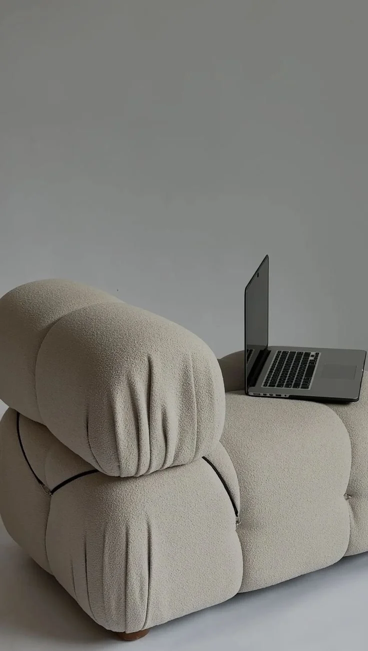 A plush beige sofa with a laptop resting on its armrest, set against a plain background.