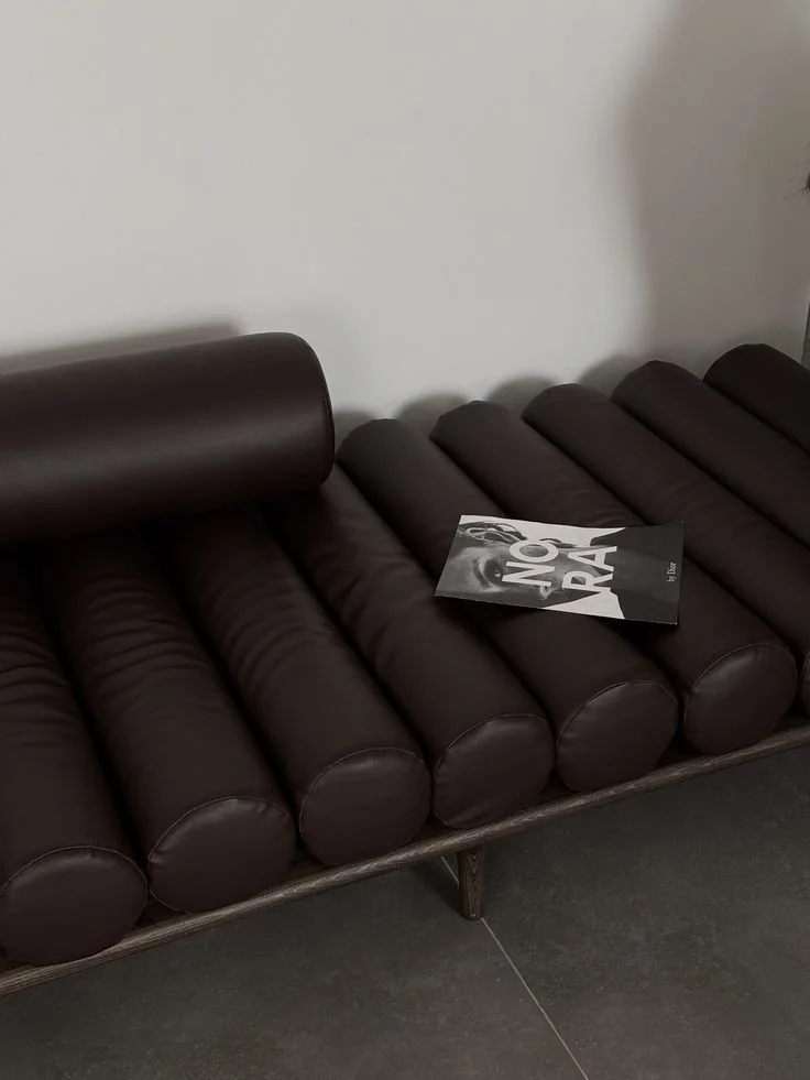 A dark brown leather bench with a cylindrical headrest and a magazine on it, positioned against a plain white wall.