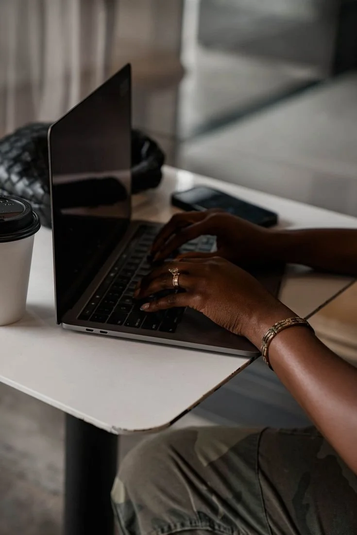 Person typing on a laptop at a white table with a coffee cup, a phone, and a quilted bag nearby.
