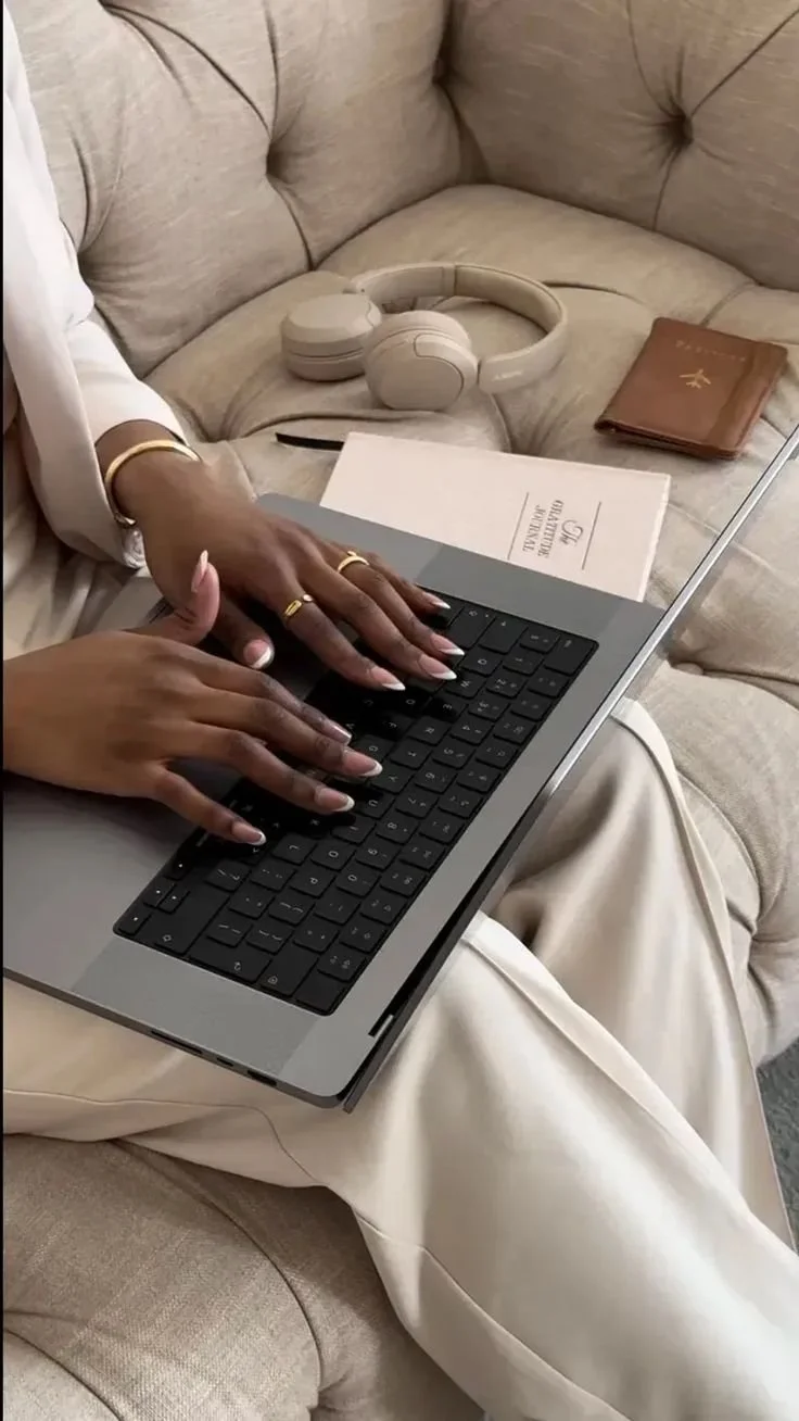 Person typing on a laptop keyboard while sitting on a beige couch with headphones, a notebook, and documents nearby.