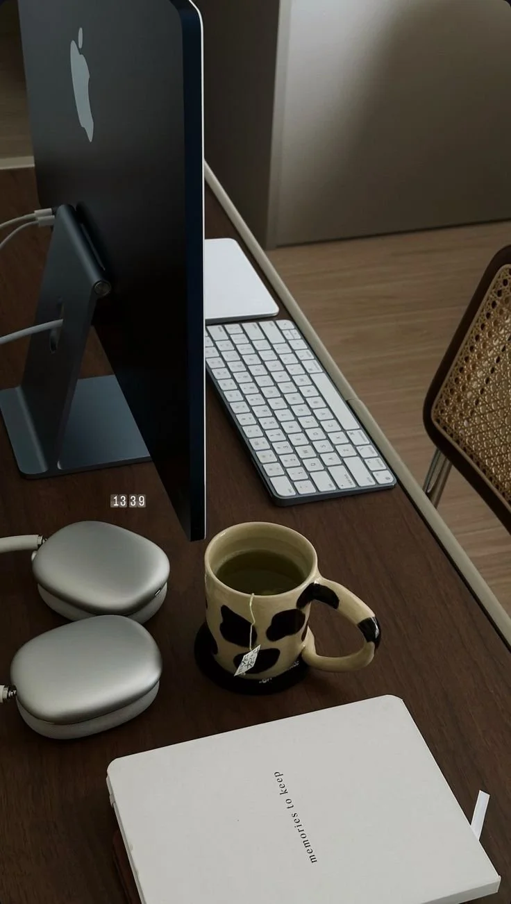 An Apple iMac computer on a desk, accompanied by a wireless keyboard, a mug of tea or coffee in a cow print mug, two closed white AirPods cases, and a notebook titled 'memories to keep'.