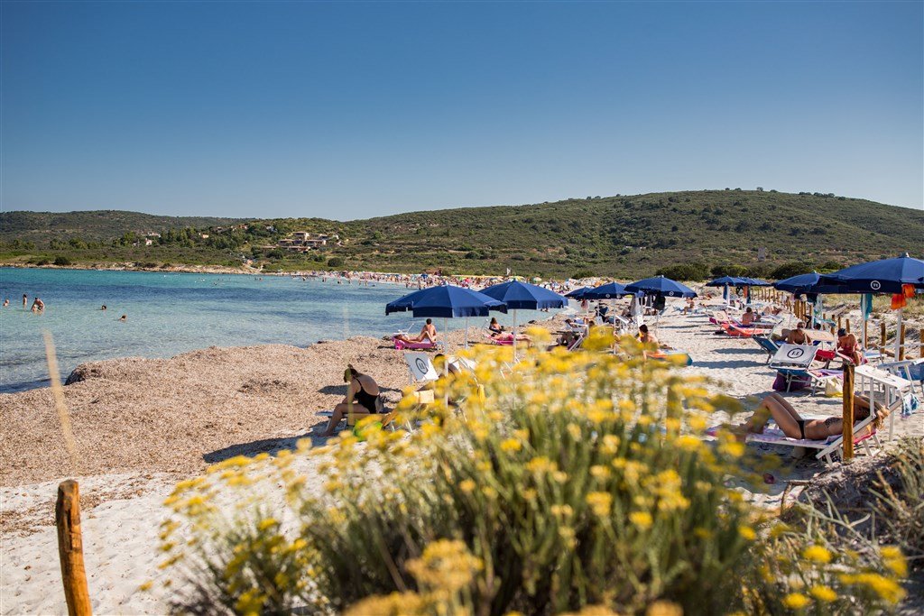 Beach scene with people relaxing on lounge chairs under blue umbrellas, swimming in clear water, and a backdrop of green hills under a sunny sky.