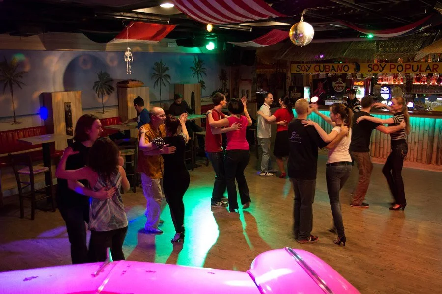 People dancing in a tropical-themed bar or nightclub with palm tree murals and colorful lighting.