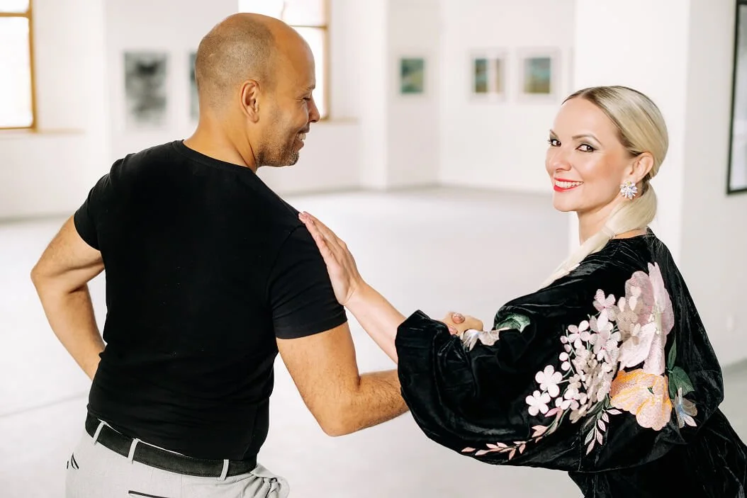 A smiling woman with platinum blonde hair and a floral kimono greets a man with a shaved head with a friendly handshake in an art gallery.