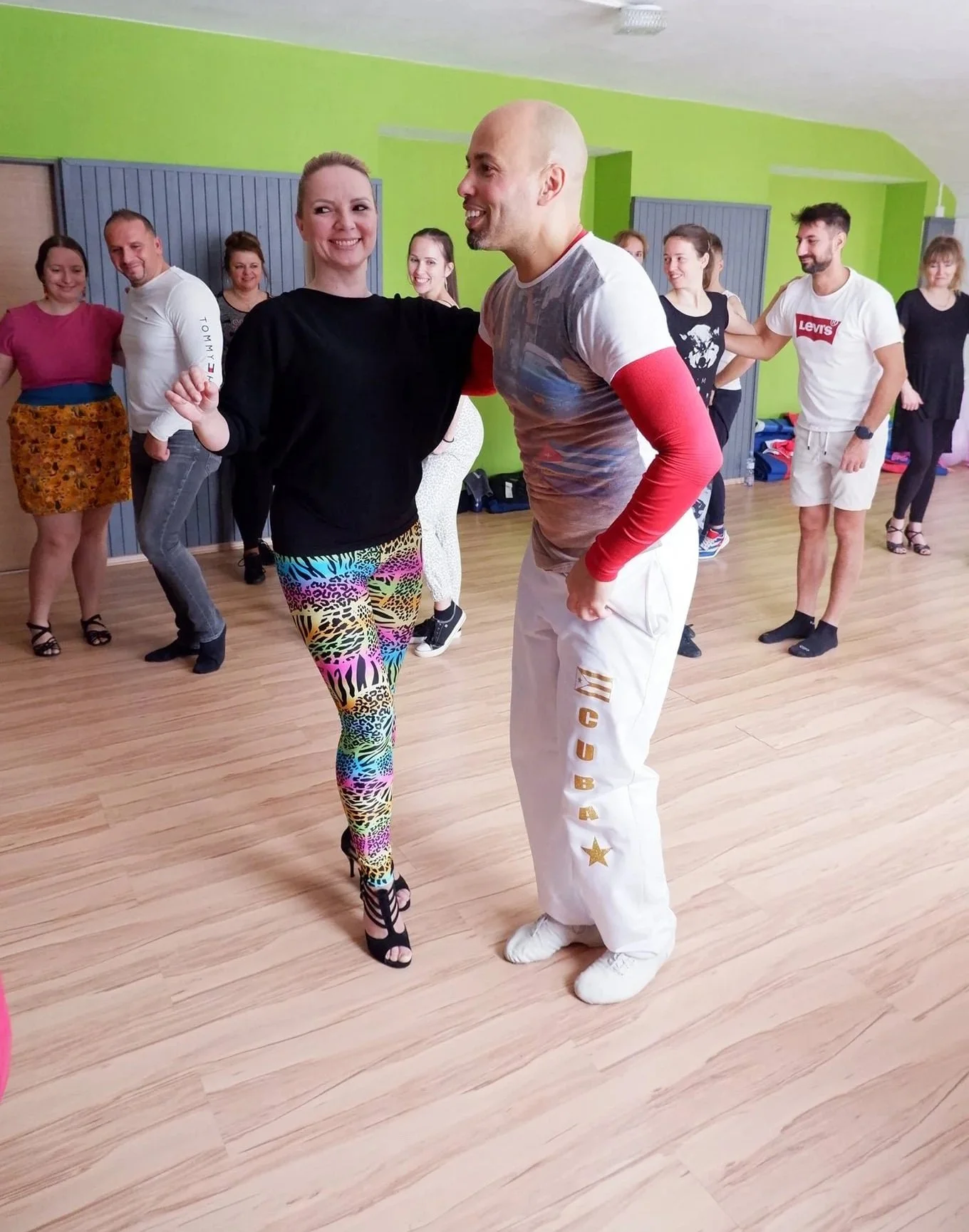 A group of people dancing together in a room with green walls, with a woman wearing colorful animal print leggings and a man in white pants with gold lettering, smiling and appearing to enjoy the dance.