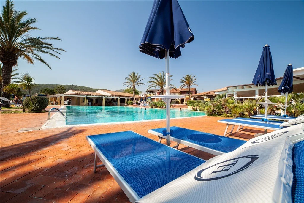 A swimming pool area with lounge chairs and umbrellas, surrounded by palm trees and lush greenery, under a clear blue sky.