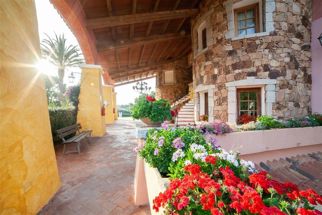 Colorful outdoor area with stone building, flowering plants, and a wooden staircase, illuminated by sunlight.