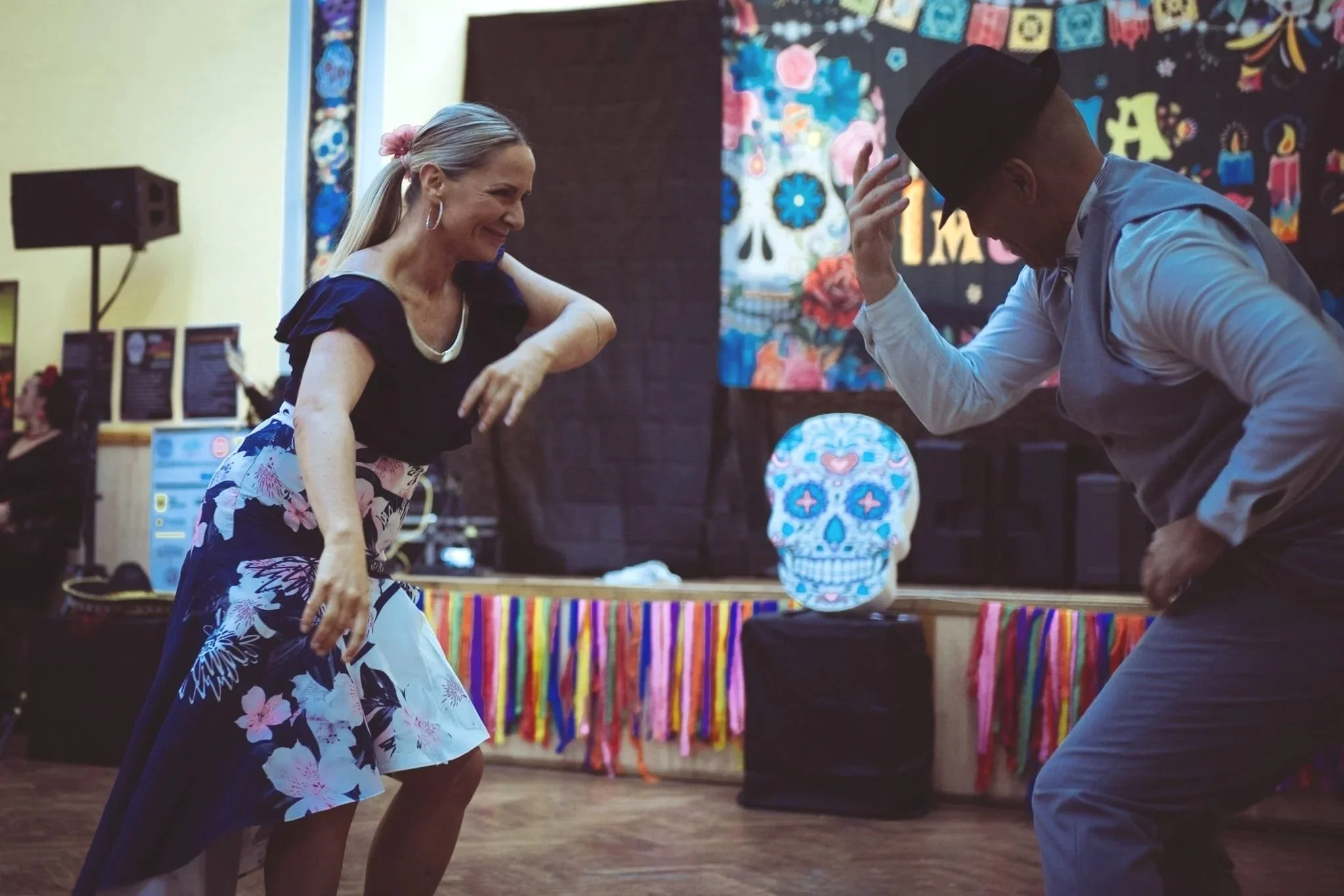 A woman and a man are dancing together at a celebration with a colorful decorated stage, a skull with floral patterns on a stand, and festive banners in the background.