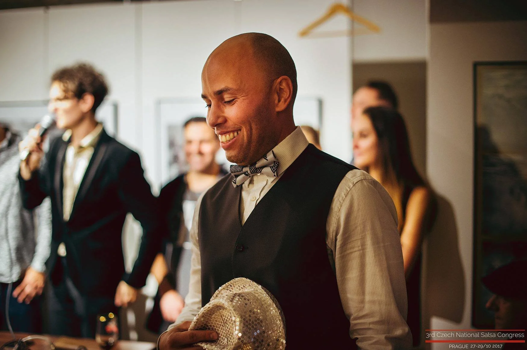 A man in a vest and bowtie smiling while holding a shiny, sequined hat, with several people in the background at an indoor event.