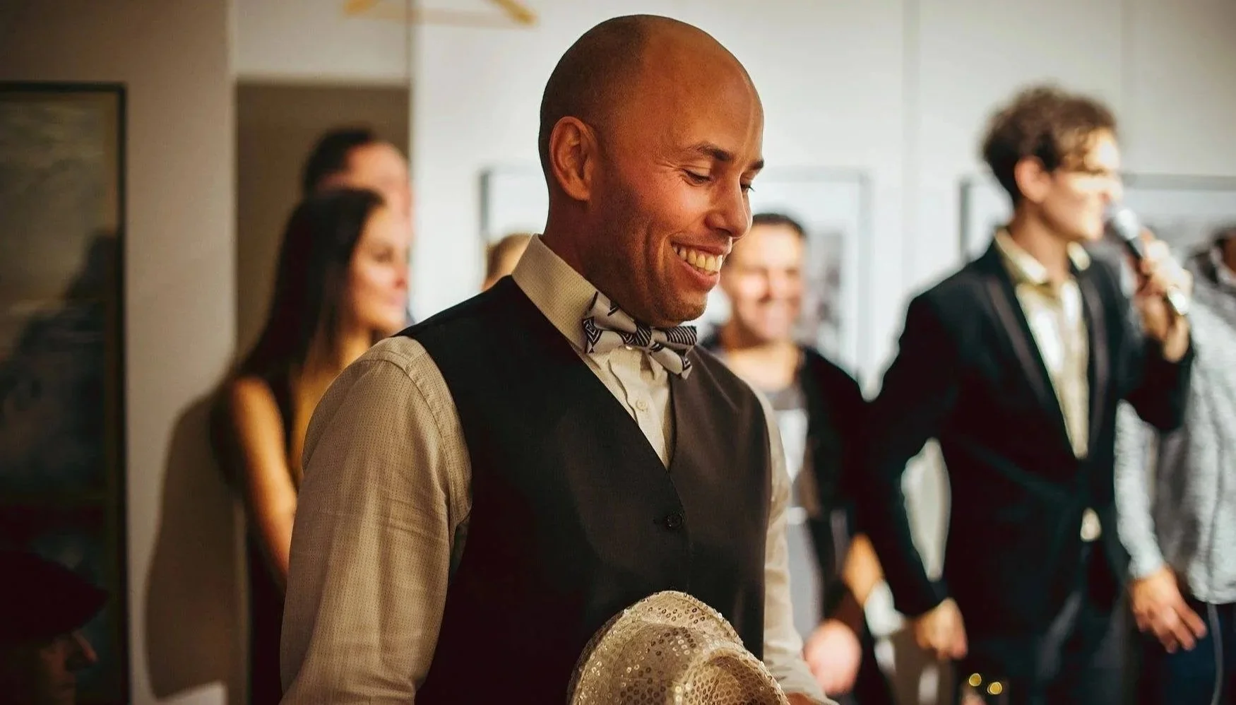 A man with a shaved head, wearing a beige shirt, black vest, and a bow tie, smiling while holding a sequined hat in a social gathering or event, with blurred people in the background.