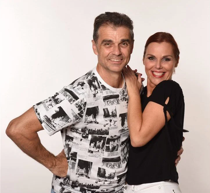 A middle-aged man and woman smiling at the camera against a plain white background. The man has short dark hair and is wearing a black and white patterned T-shirt. The woman has red hair and is dressed in a black top with short sleeves. They are standing close together, with the woman's hand on the man's shoulder and the man's arm around her waist.
